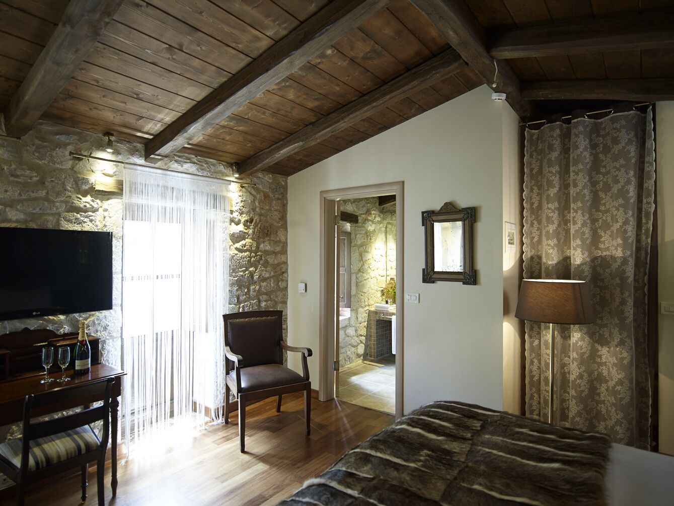 Een knus ingerichte kamer met houten plafond, stenen muren, een bed, een televisie, een stoel en een lamp.