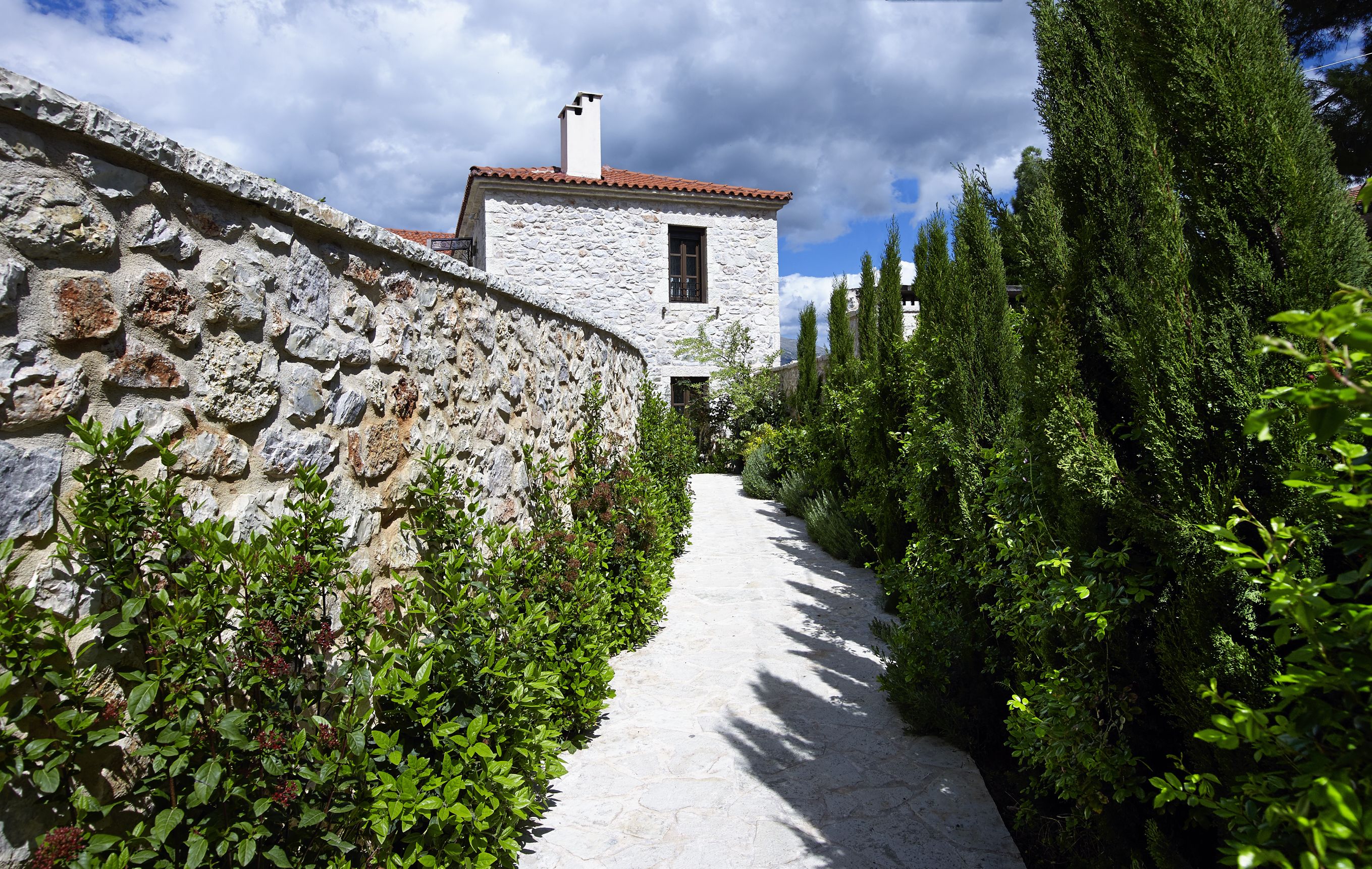 Un sendero de piedra que conduce a un edificio blanco con techo de tejas rojas, rodeado de follaje y árboles altos.