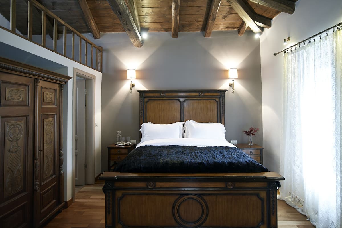 A bedroom with a wooden bed, two wall-mounted lamps, a nightstand, and a window with sheer curtains.