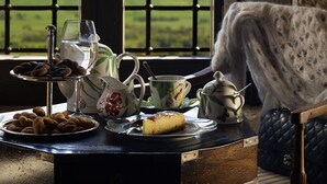 A table with a tea set, a plate of pastries, and a slice of cake, with a scenic view of mountains through a window. - Villa Vager (Tripoli)