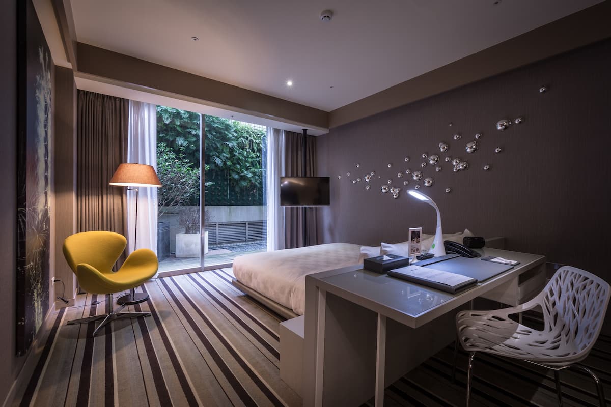 Quarto de hotel moderno com cama, escrivaninha, cadeira, televisão e janela com cortinas.