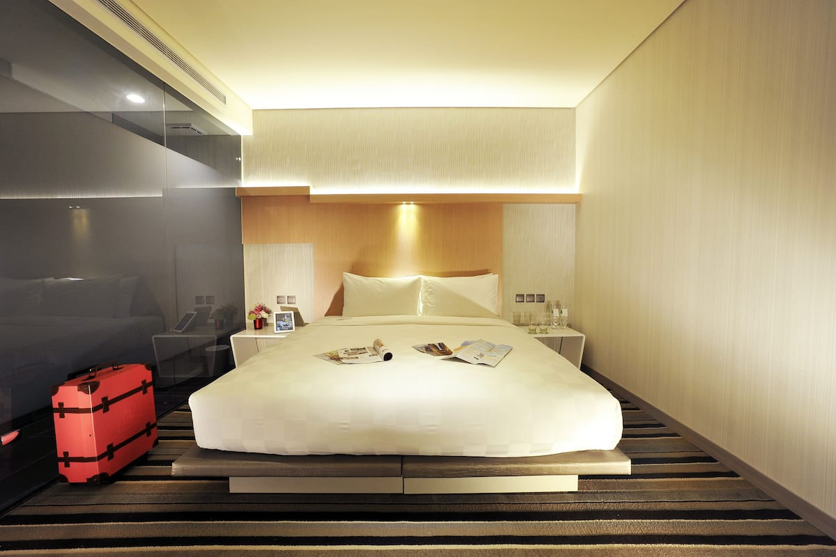 Quarto de hotel com cama, mala vermelha, mesa de cabeceira e abajur.