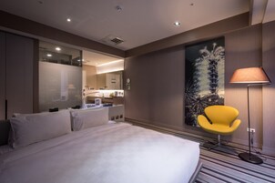 Een moderne hotelkamer met een groot bed, een felgele stoel, een grote spiegel en uitzicht op de keuken en het balkon.