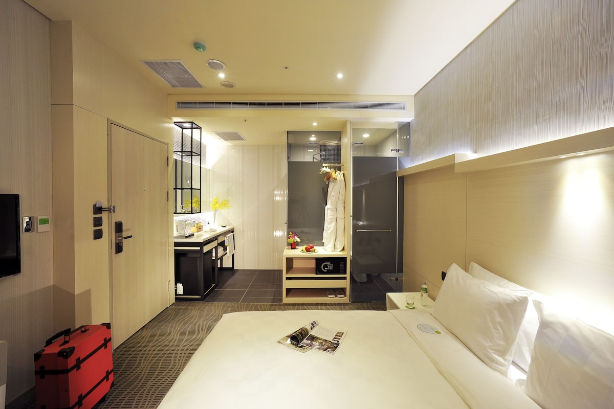 Quarto de hotel com uma cama, uma escrivaninha, um chuveiro e uma mala vermelha.