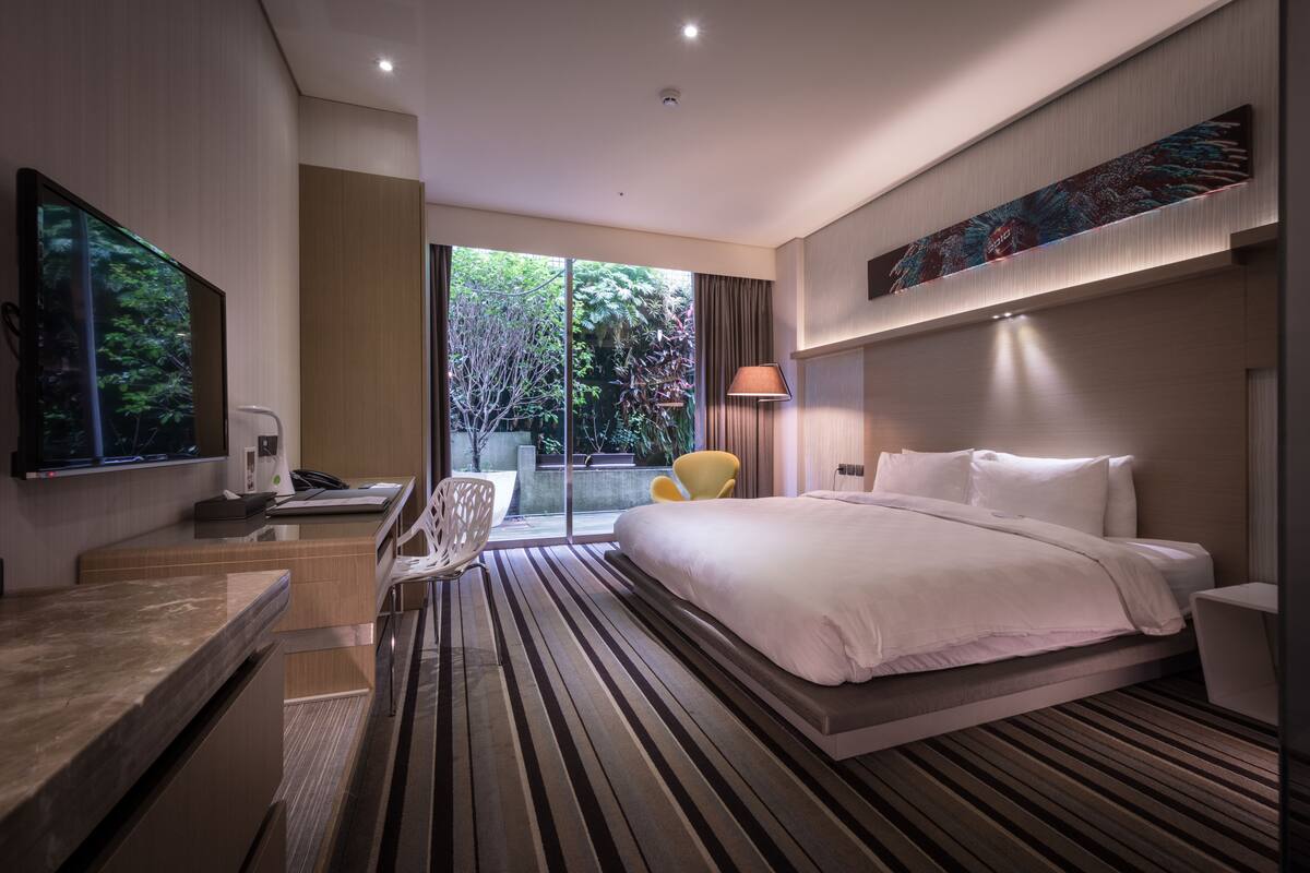 Quarto de hotel moderno com uma cama grande, uma mesa de trabalho e uma cadeira. Há uma varanda com vista para árvores.