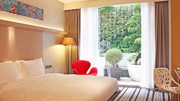A hotel room with a large bed, a red chair, a white chair, a lamp, and a view of greenery outside.