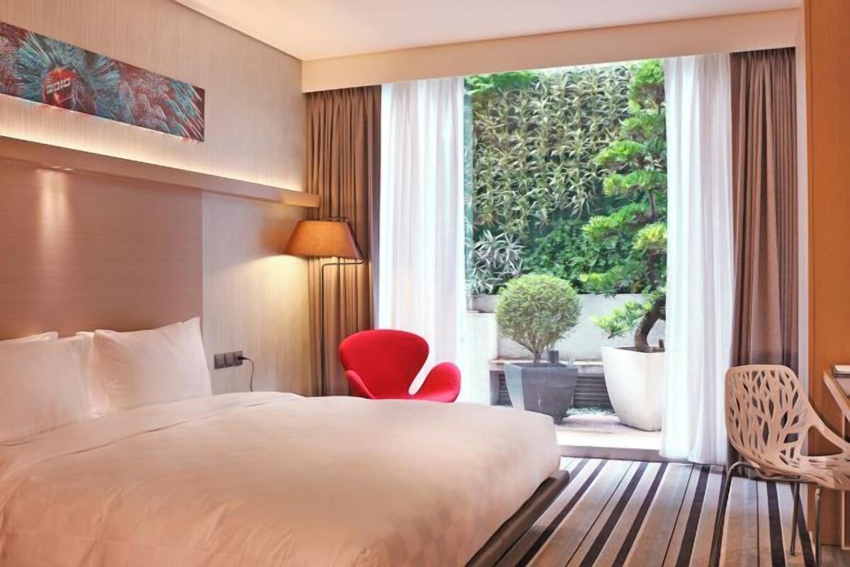 A hotel room with a large bed, a red chair, a potted plant, and a view of greenery outside.