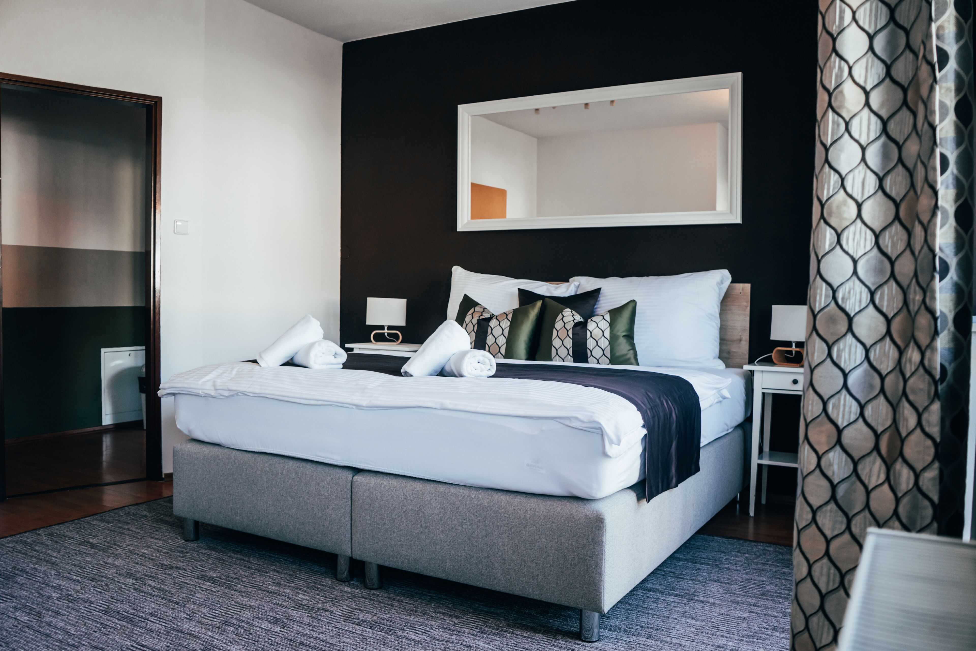 Moderni hotellihuone, jossa on suuri sänky, yöpöydät, peili ja kuviollinen verho.
