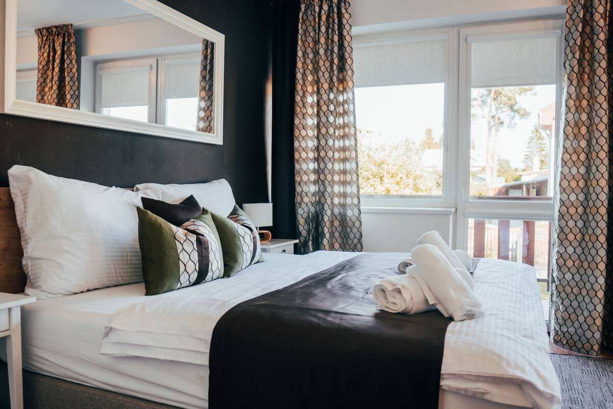 A neatly made bed with white and black bedding, a black headboard, and a window with patterned curtains.