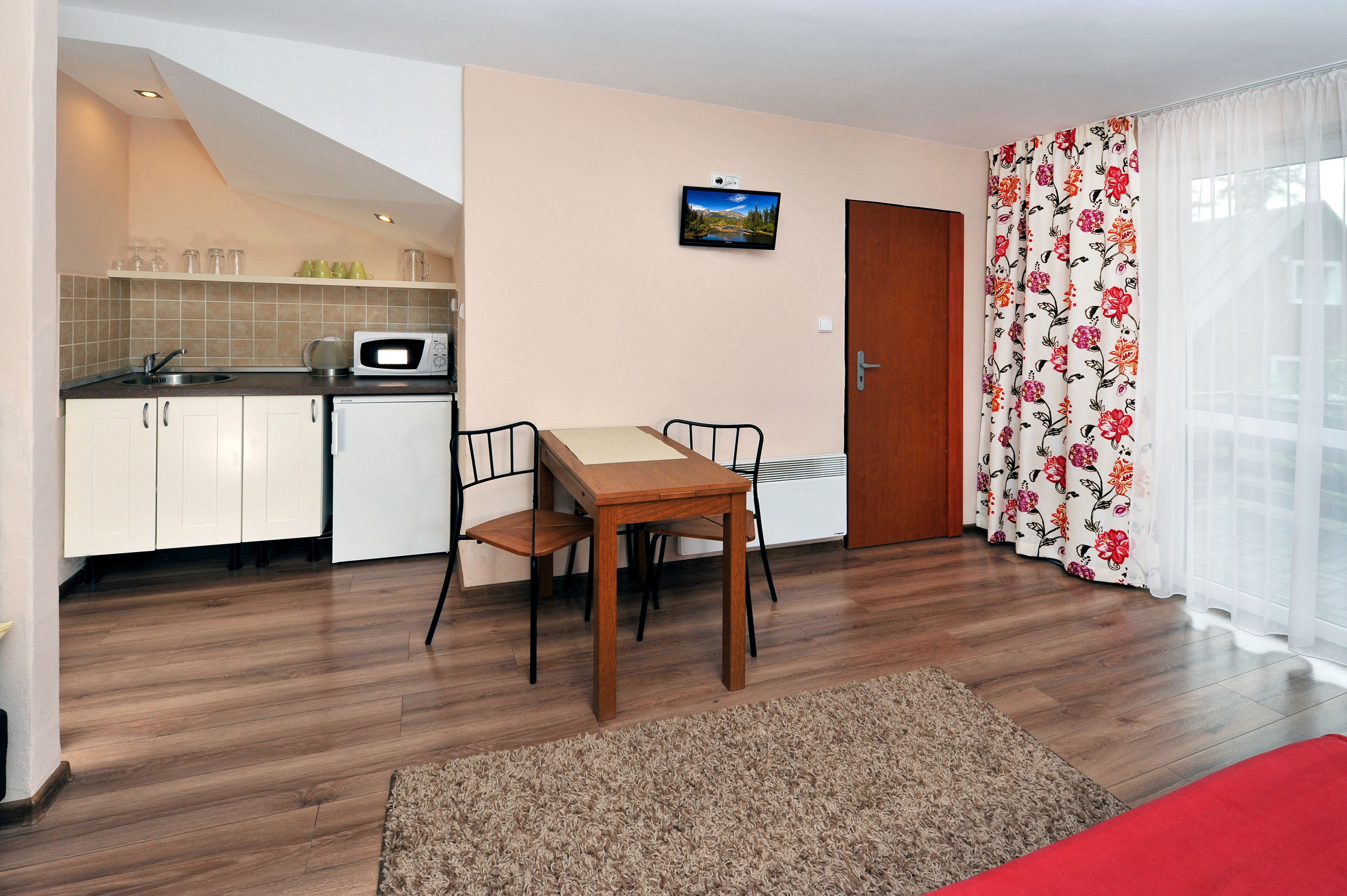 A compact kitchen with white cabinets, a microwave, and a small sink. There is a dining area with a wooden table and chairs. The room has a wooden floor and a red bed in the corner.