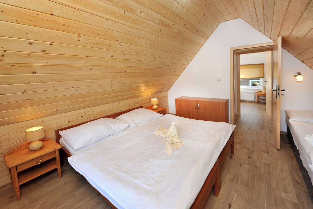 A bedroom with a wooden ceiling, a bed with white linens, a nightstand with a lamp, and a wooden wardrobe.