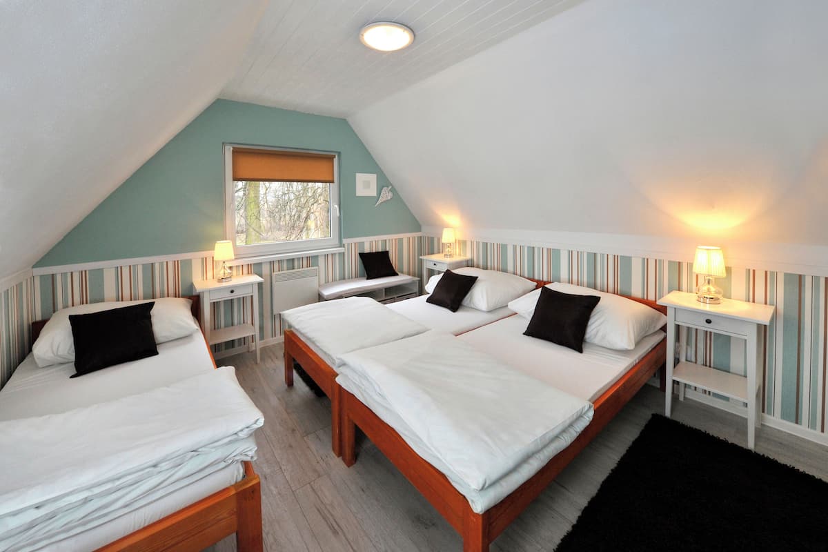 A room with three beds, each with white bedding and black pillows, wooden headboards, and striped wallpaper. There are bedside tables with lamps and a window with a blind.