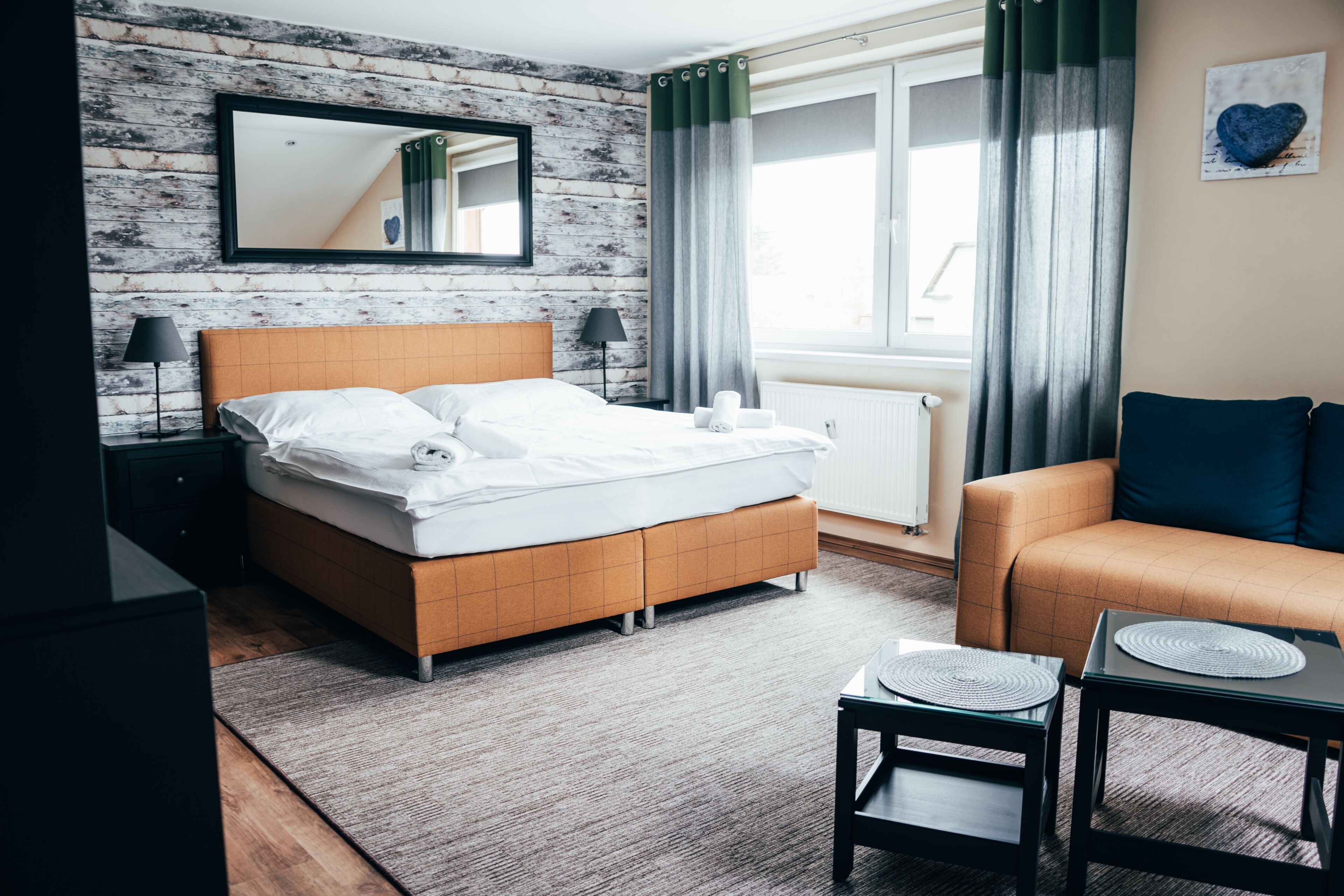 Une chambre d’hôtel moderne avec un mur en bois, un grand lit avec du linge de lit blanc, un canapé, une petite table et une fenêtre avec des rideaux.