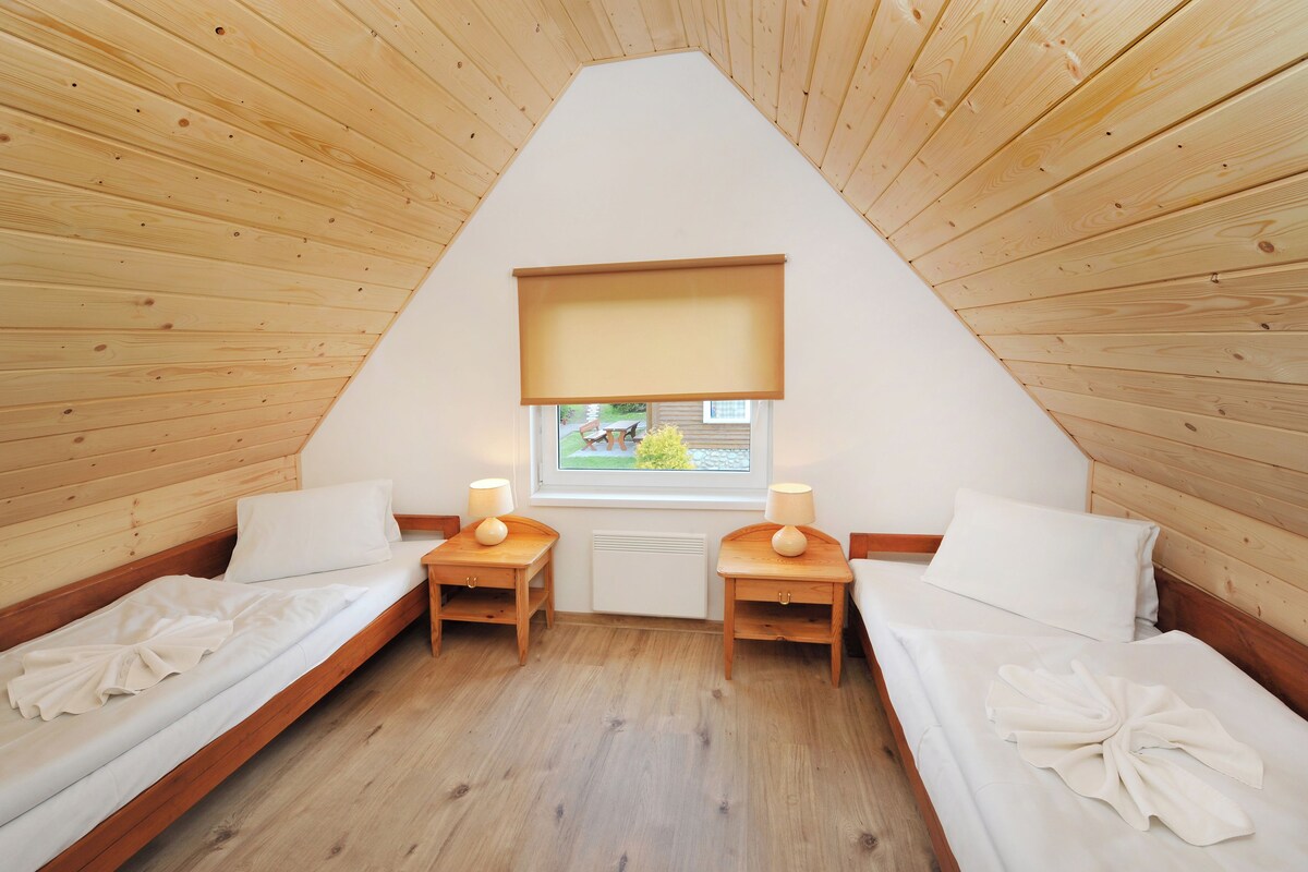 A small, cozy room with wooden walls and a sloped ceiling. There are two single beds with white linens and matching nightstands with lamps. A window with a view of a garden is visible.