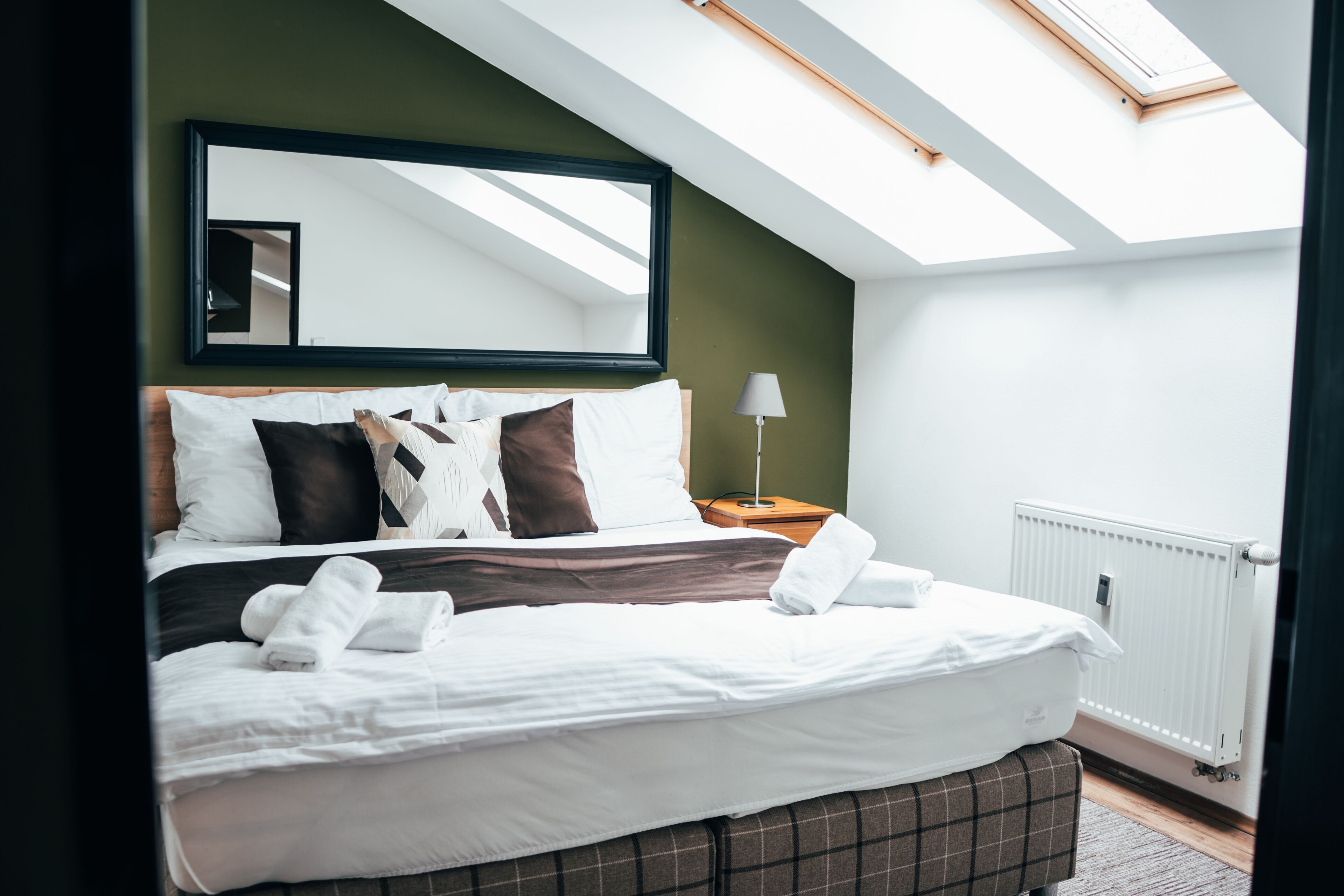 Une chambre à coucher moderne avec un puits de lumière, un grand lit avec du linge de lit blanc, une table de chevet avec une lampe et un radiateur.