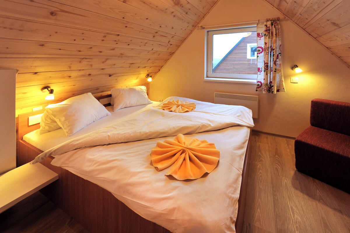 A cozy bedroom with a sloped ceiling, a bed with white linens, two bedside lamps, and a window with curtains.