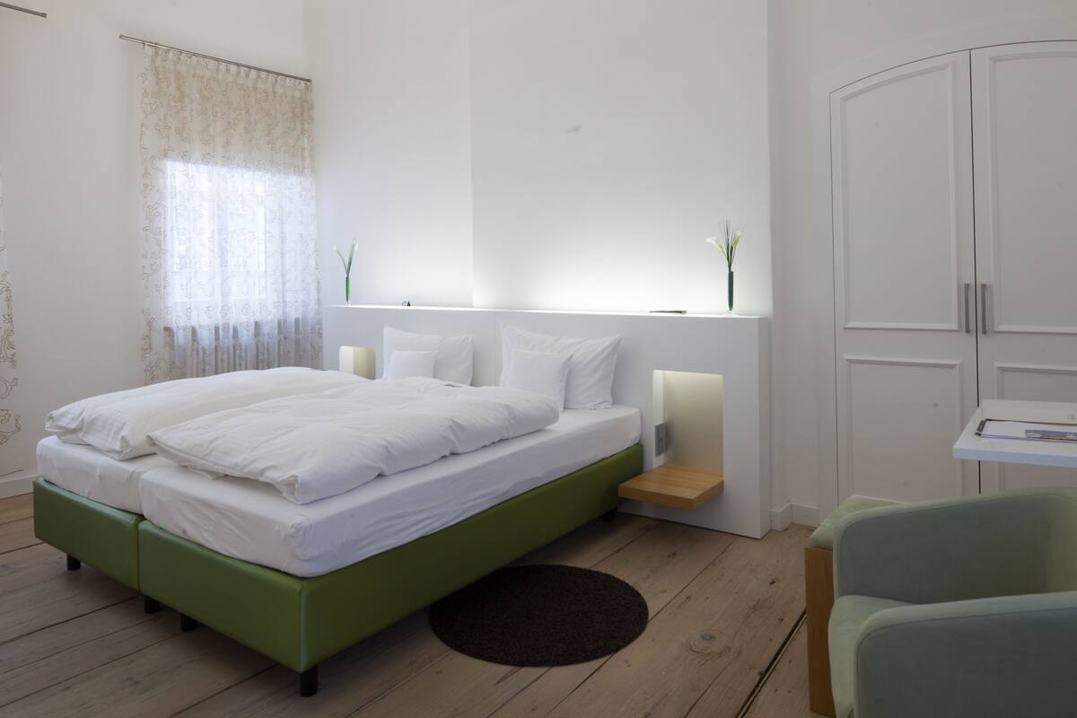 Un dormitorio moderno con un cabecero verde, ropa de cama blanca, una mesita de noche con lámpara y un armario.