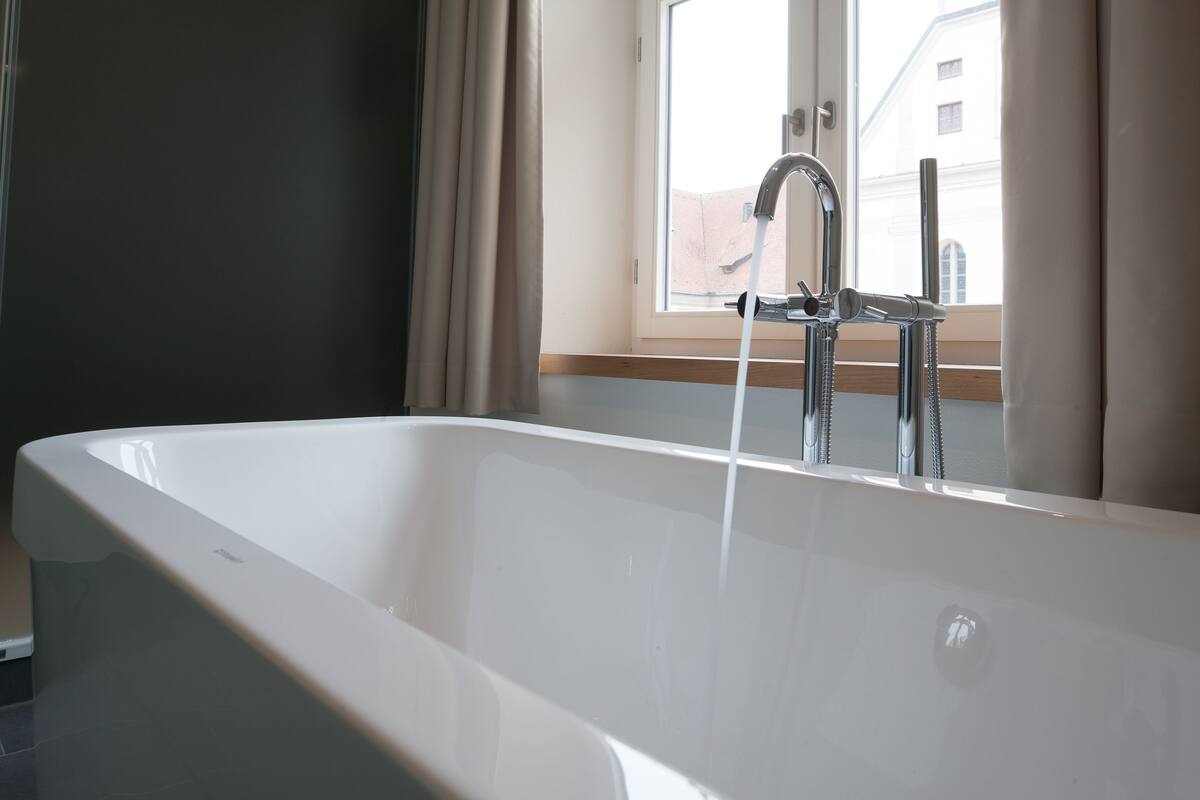 Un baño moderno con una bañera independiente, una ventana con cortinas y vistas a edificios.