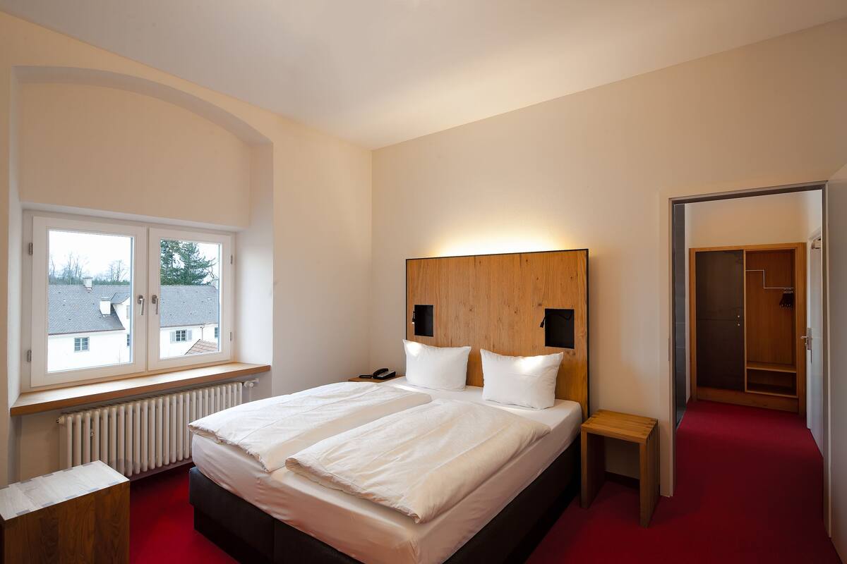 Un dormitorio con una cama grande, cabecera de madera, una mesita de noche y una ventana con vista a edificios.
