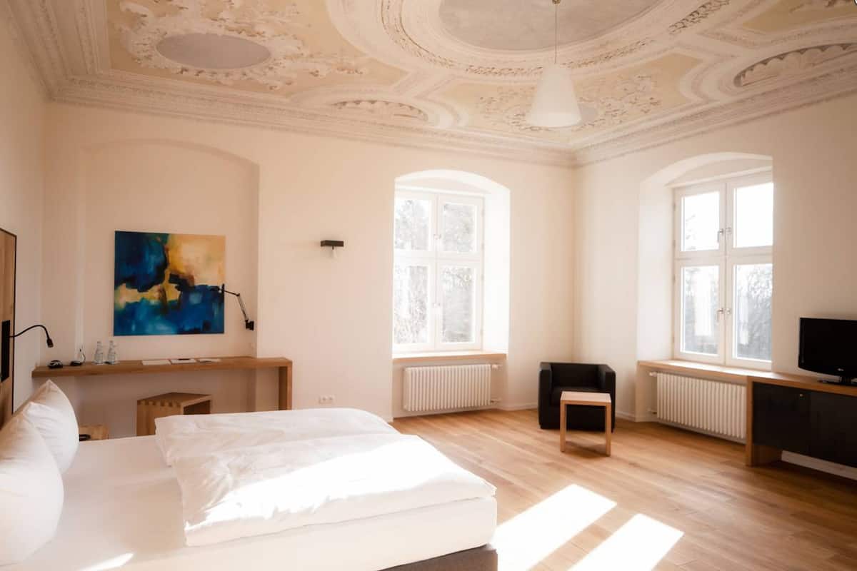 Un dormitorio amplio con una cama grande, un escritorio, una silla y una televisión. Hay dos ventanas que ofrecen luz natural y vistas al exterior.