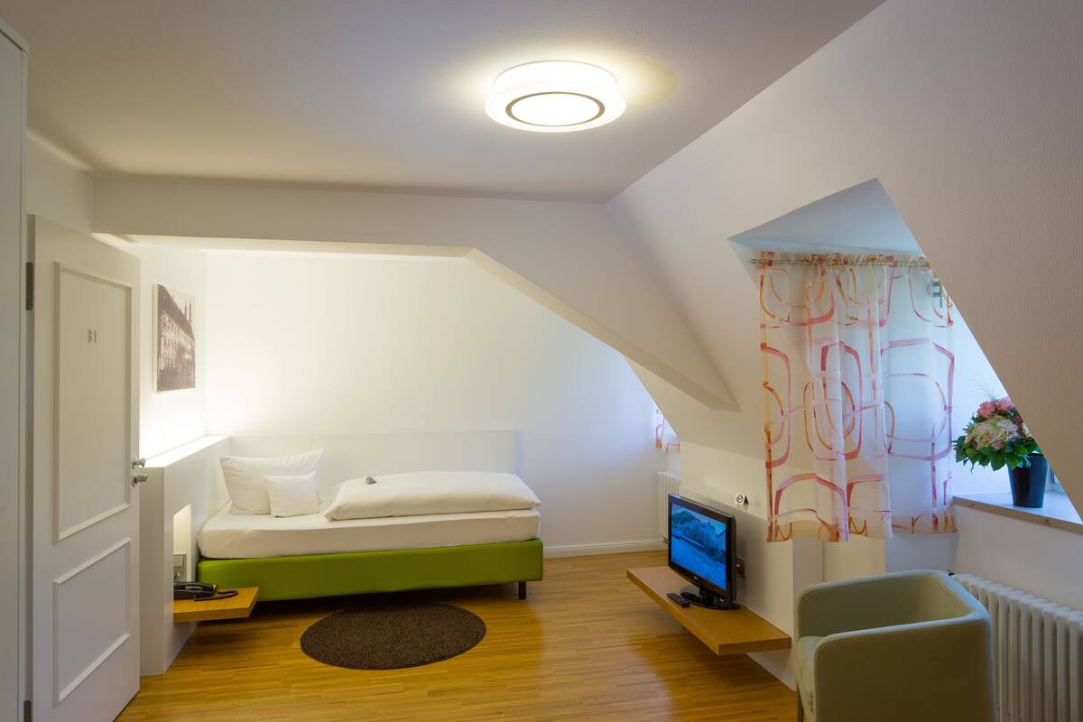 Un dormitorio pequeño y moderno con una cama verde, un televisor, un escritorio y una ventana con cortina.