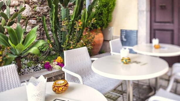A patio area with white round tables and chairs, a stone wall with cacti and other plants, and a door in the background. - Porta del Tocco Design Rooms (Taormina)