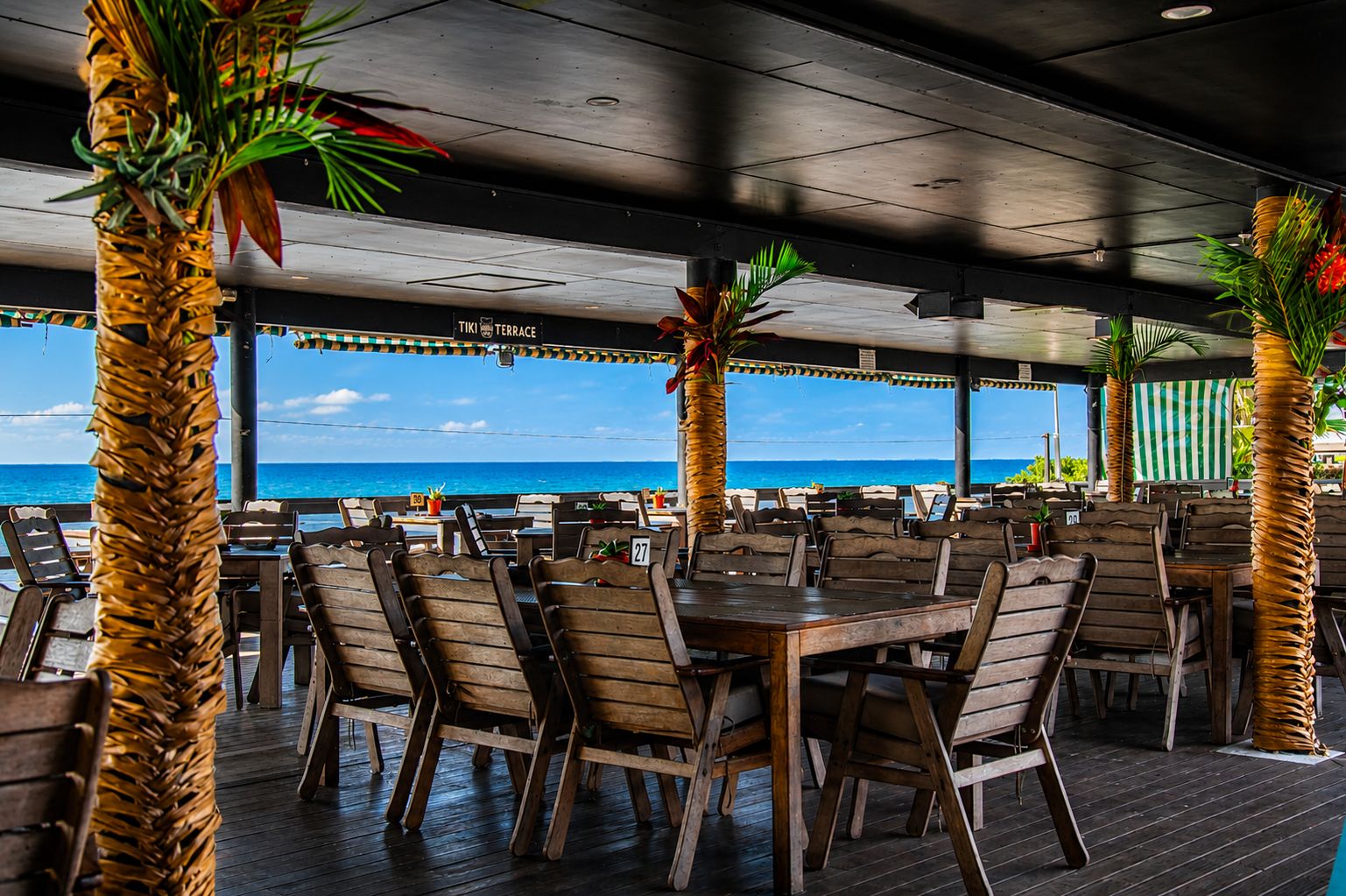 Un espace de restauration en plein air avec des tables et des chaises en bois, entouré de palmiers et offrant une vue sur la plage.
