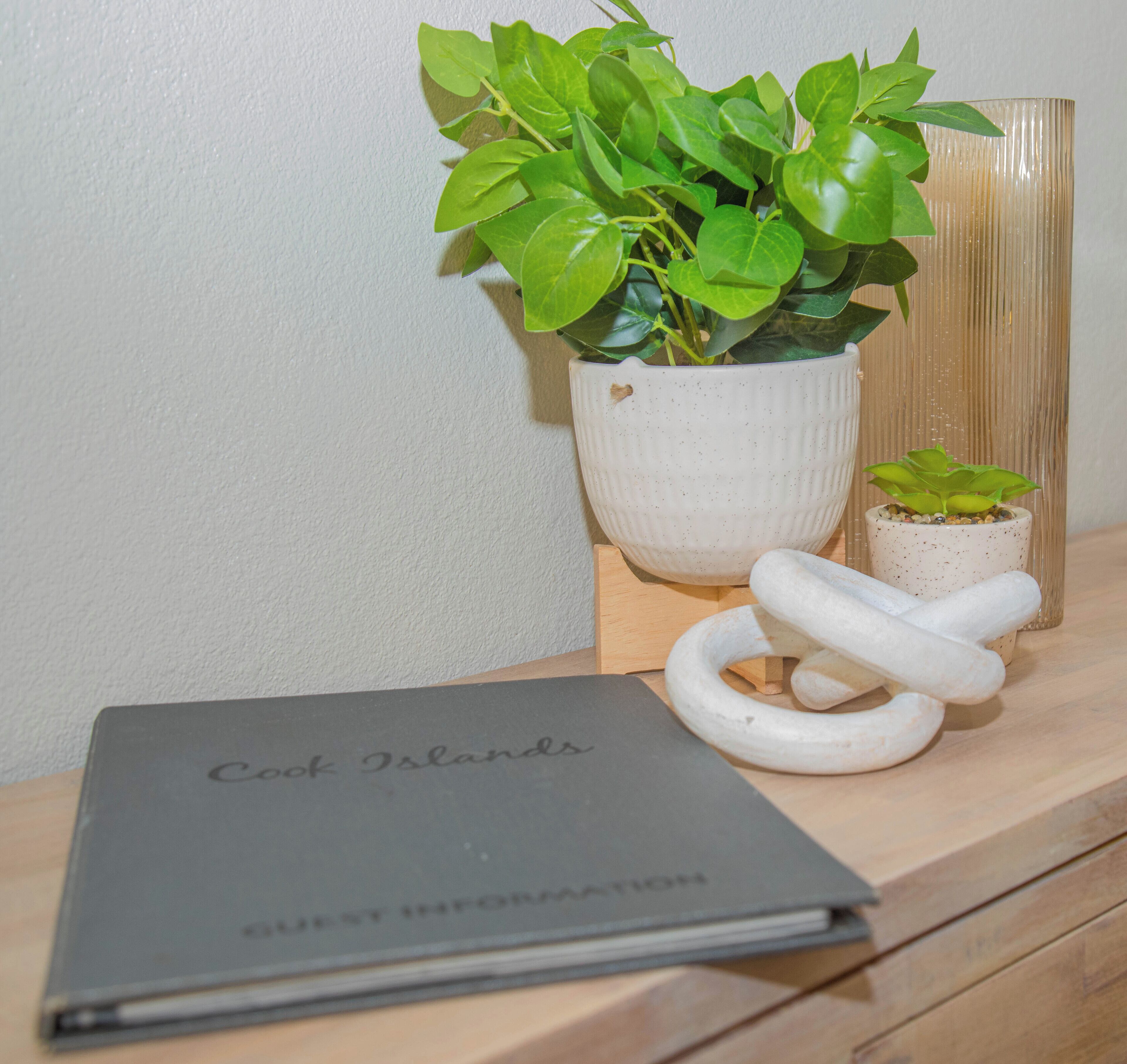 A book titled "Cook Islands Guest Information" on a wooden surface, a white pot with a plant, and a small white sculpture.