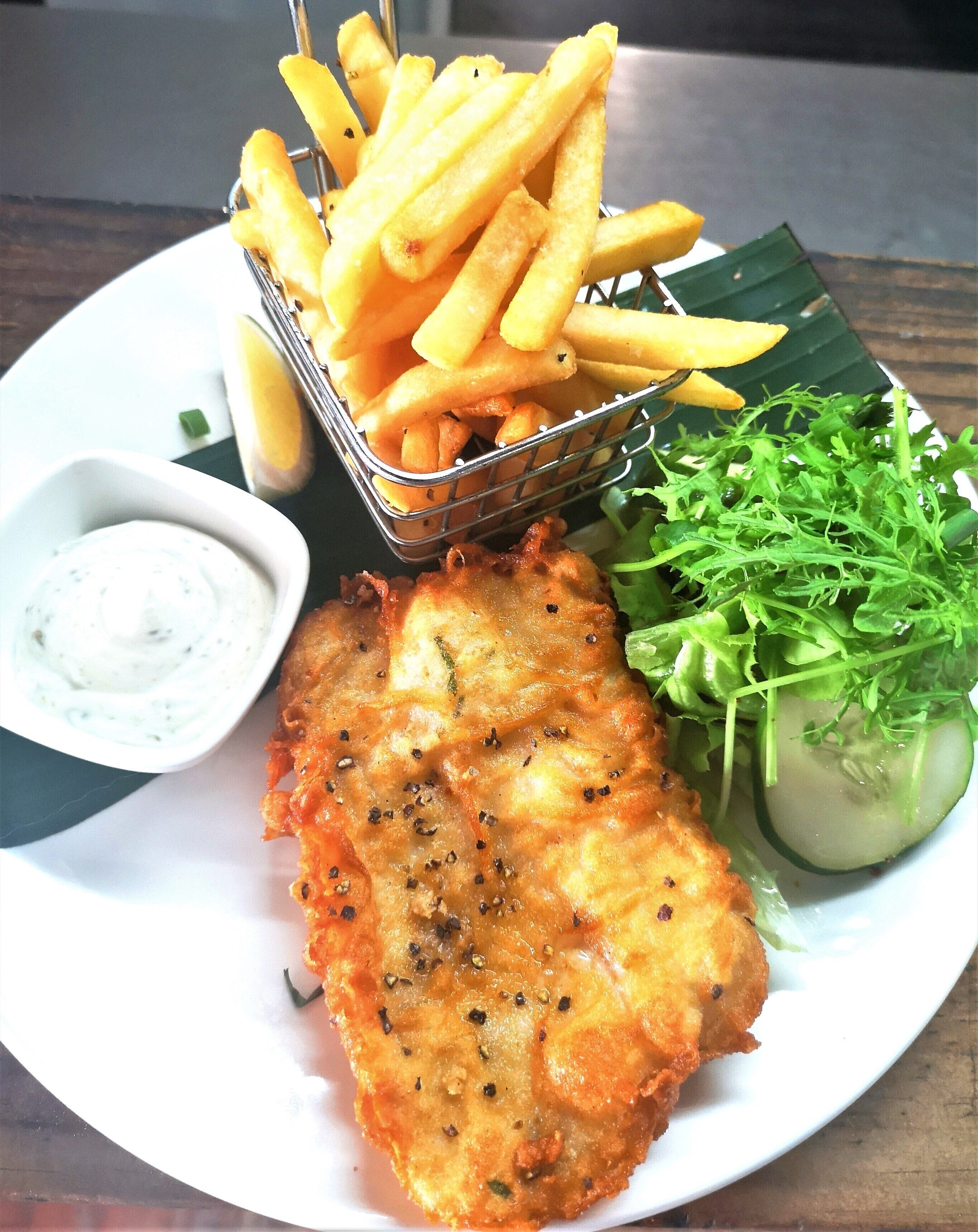 A plate with a breaded fish fillet, French fries, a small bowl of tartar sauce, a cucumber slice, and a side salad.