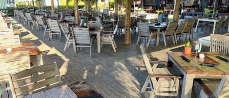 Un área de comedor al aire libre con mesas y sillas de madera, rodeada de follaje y un corredor cubierto.