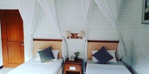 Quarto com duas camas, paredes de tijolos brancos, uma porta de madeira e uma mesa de cabeceira com abajur.