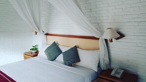 A neatly made bed with a white headboard, two pillows, a bedside lamp, and a small table with a plant. - Puri Padi Hotel (Ubud)