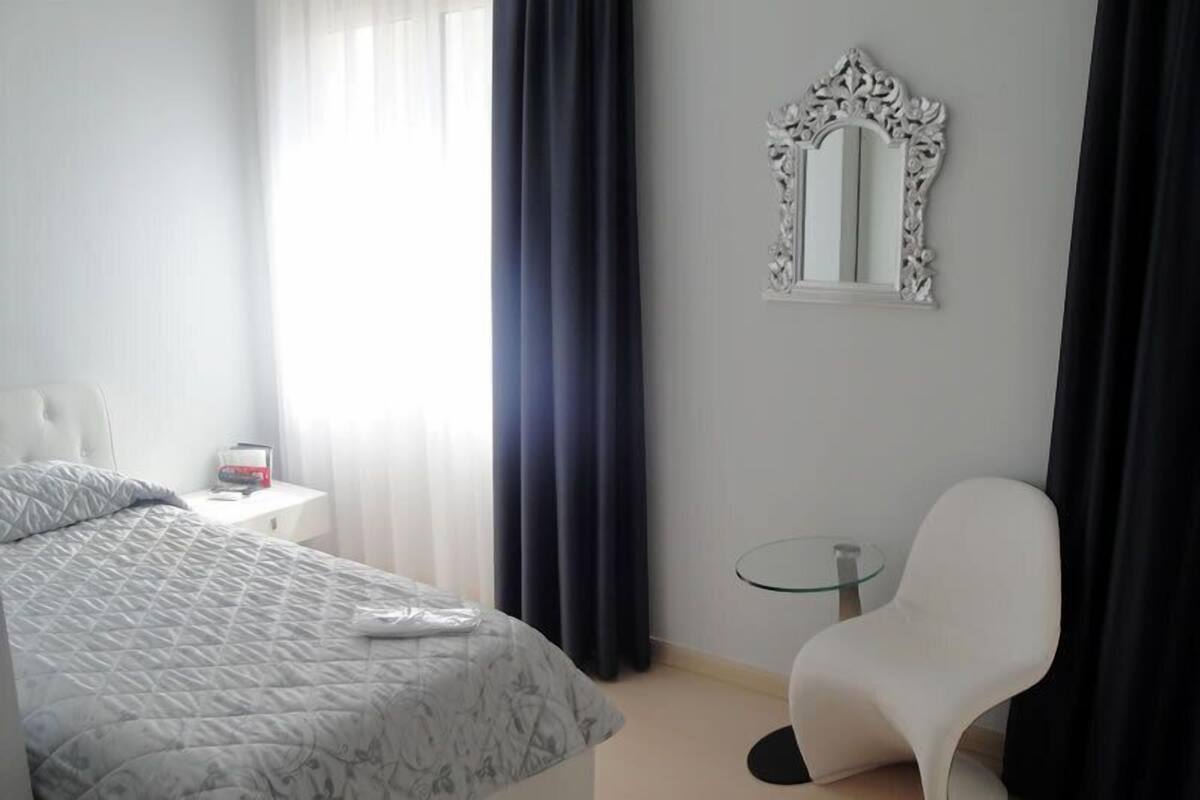A hotel room with a bed, a chair, a small table, a mirror, and a window with curtains.