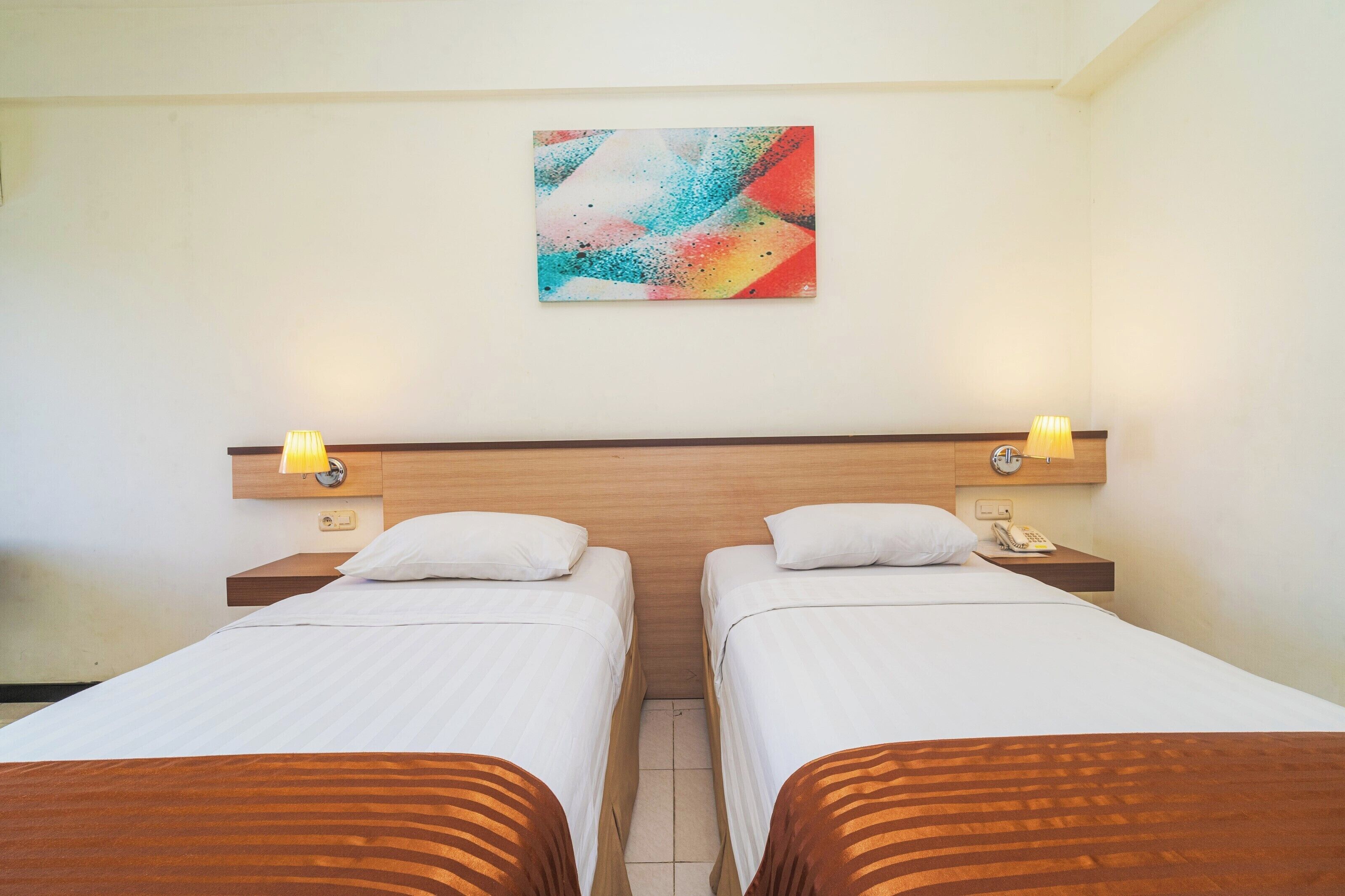Habitación de hotel con dos camas individuales, cabecera de madera, lámparas de noche, teléfono y un cuadro abstracto de colores en la pared.