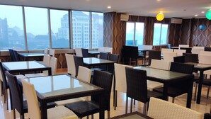 Zona pranzo moderna con tavoli in vetro e sedie in vimini, ampie finestre con vista sulla città e un design murale decorativo.