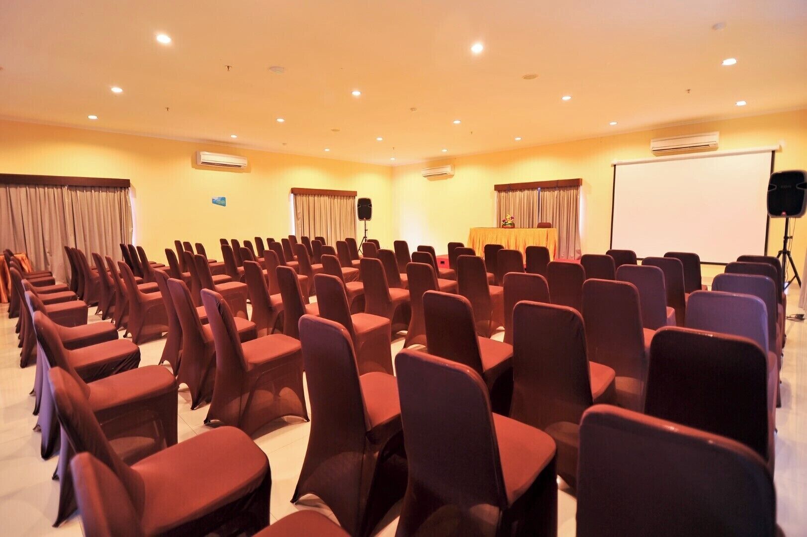 A conference room with rows of chairs, a projector screen, and an air conditioning unit on the wall.