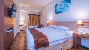 Una camera d'albergo con un letto grande, una TV fissata al muro, un quadro di una rosa blu e un comodino con una lampada.