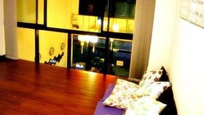A room with a wooden floor, a bed with a patterned bedspread, a window with a view of the outside, and a wall-mounted TV. - de Talak (Bangkok)