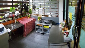 Eine kompakte Küche mit Spüle, Herd und Tisch, bedeckt mit einer rot-weiß karierten Tischdecke. An einem Gestell über der Spüle hängen Töpfe und Pfannen, und ein Schild weist an, die Küche nach Gebrauch zu reinigen.