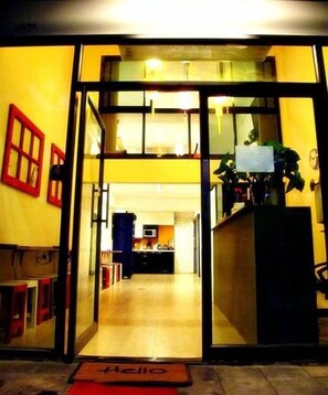 Entrada de un hotel con puerta de vidrio, una recepción y una cocina visible al fondo.