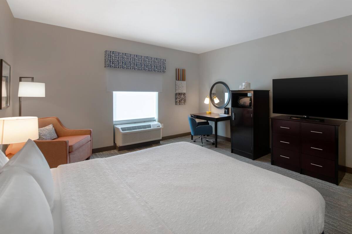 Une chambre d’hôtel avec un grand lit, un bureau avec une chaise, une télévision et une fenêtre avec des stores.