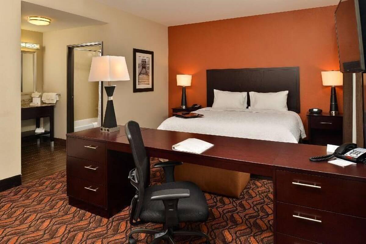 Une chambre d’hôtel avec un lit, un bureau, une chaise, des lampes, et une salle de bain visible au fond.