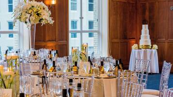 A banquet hall with a long table set for a formal event, featuring clear glass chairs, a tiered white cake, and floral centerpieces.