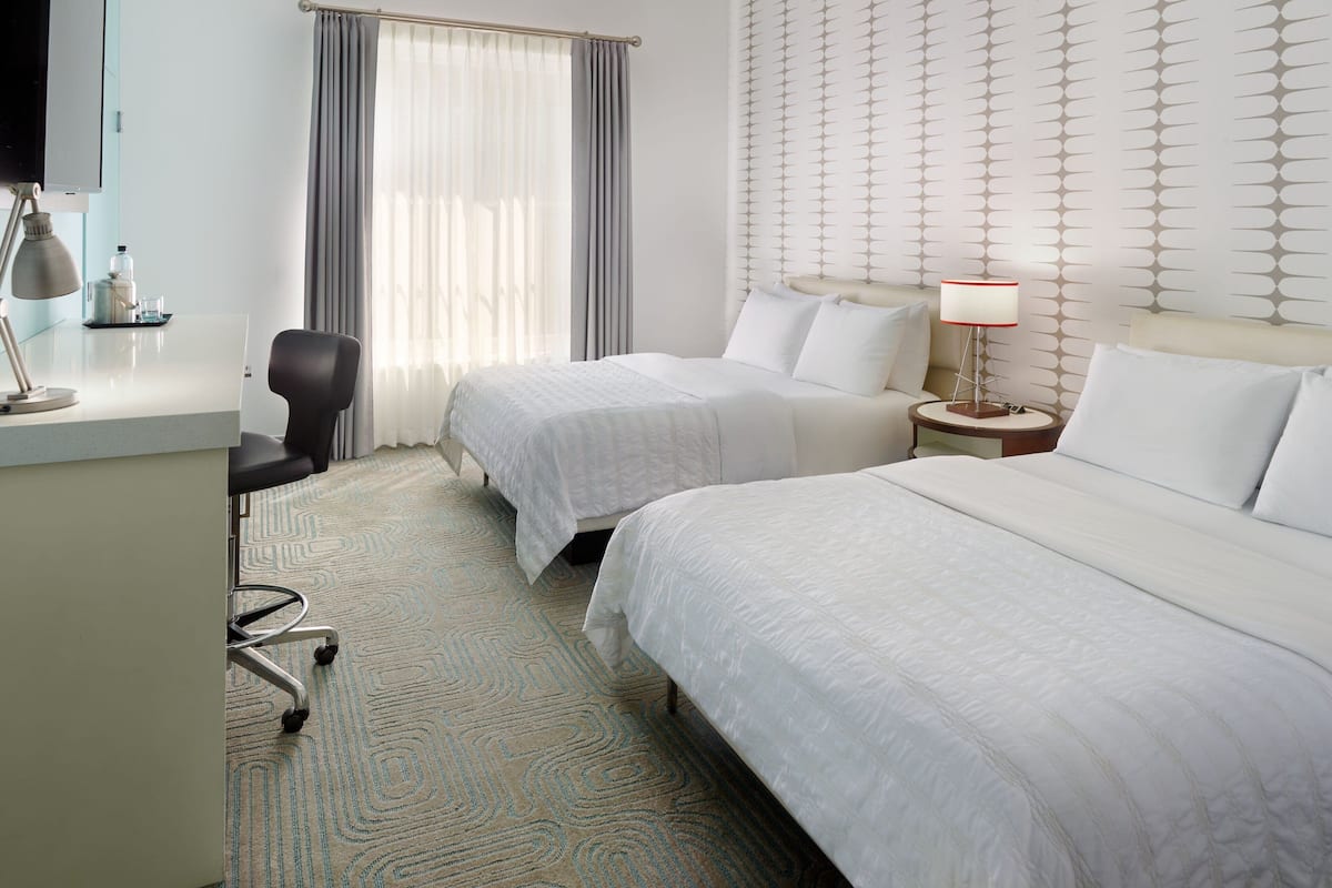 A hotel room with two beds, a desk, a chair, a lamp, and a window with curtains.