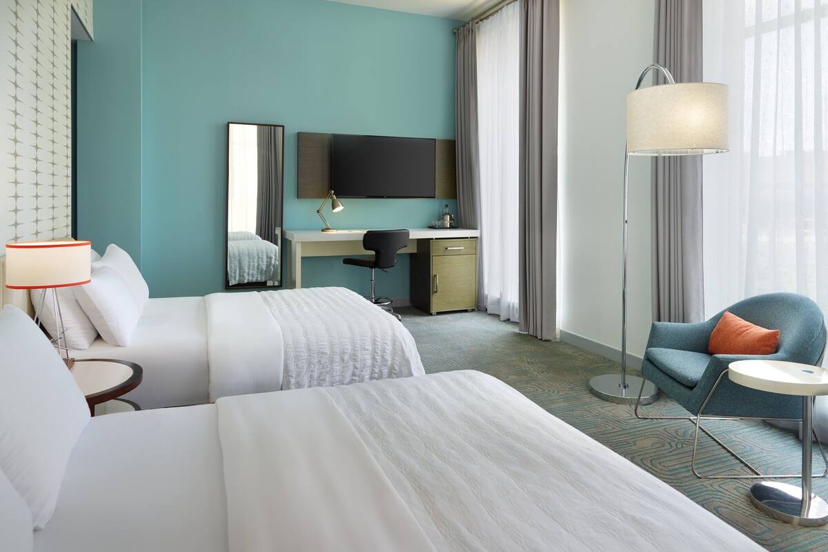 A hotel room with two beds, a desk, a chair, a TV, and a lamp.