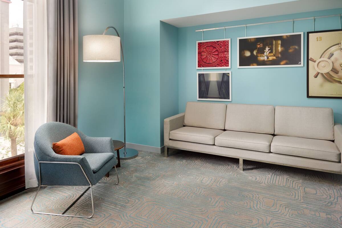 A room with a blue wall, a beige sofa, a blue armchair with an orange pillow, a floor lamp, and patterned carpet.