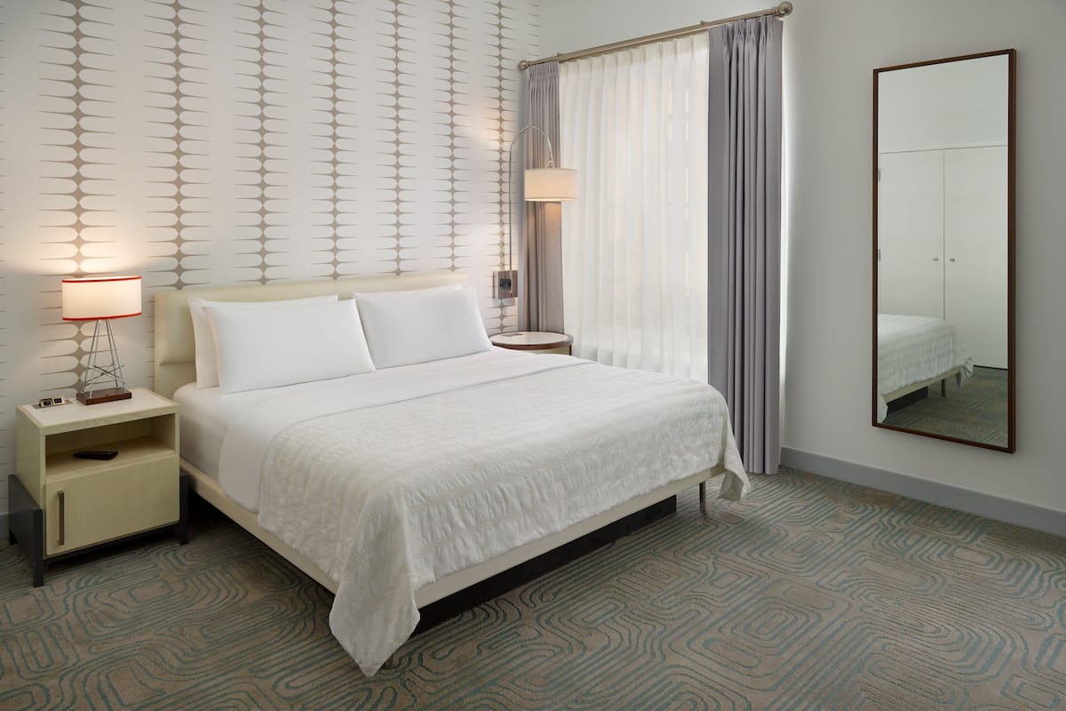 A neatly arranged hotel room with a bed, bedside tables, a lamp, a mirror, and patterned carpet.