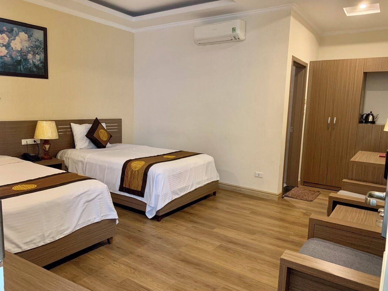 A hotel room with two beds, a wooden wardrobe, a desk, and an air conditioning unit.
