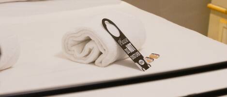 A neatly made bed with a rolled white towel and a black tag, a white pillow, and a small shelf with a bouquet of flowers.