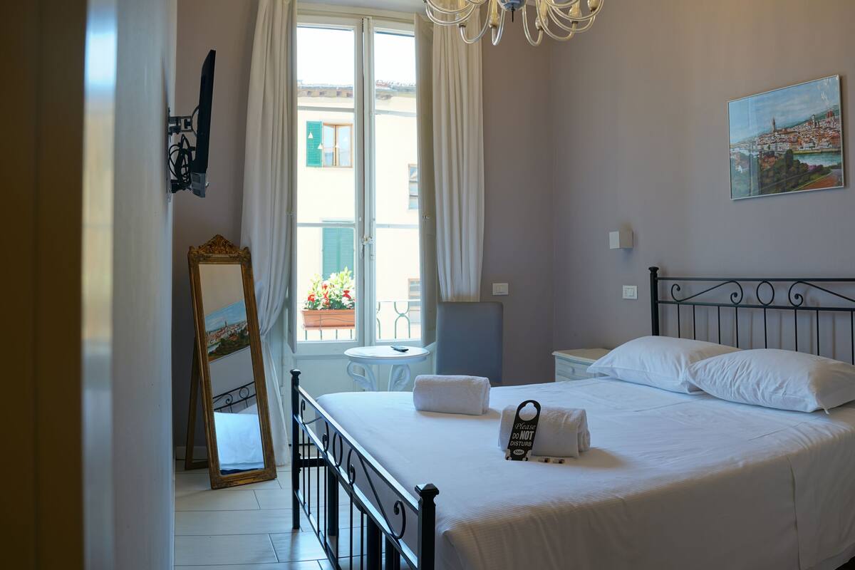 A bedroom with a bed, bedside tables, a nightstand, a mirror, a window with curtains, and a painting on the wall.
